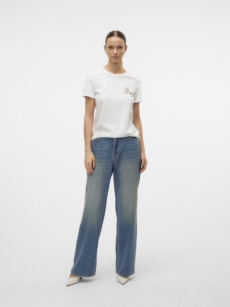 Μοντέλο φοράει VERO MODA λευκό t-shirt με τύπωμα GOLD GLITTER STILLETO, συνδυασμένο με τζιν παντελόνι